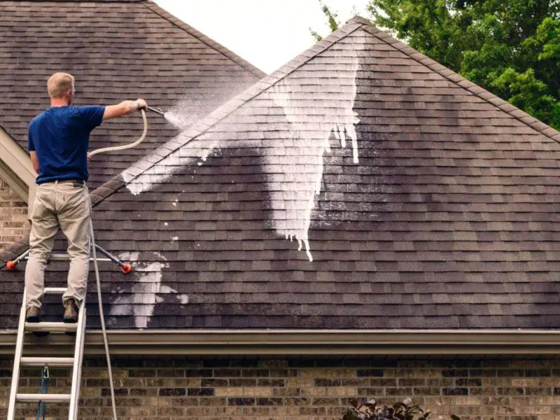 A Supreme Clean field technician soft washing a roof while standing on a ladder